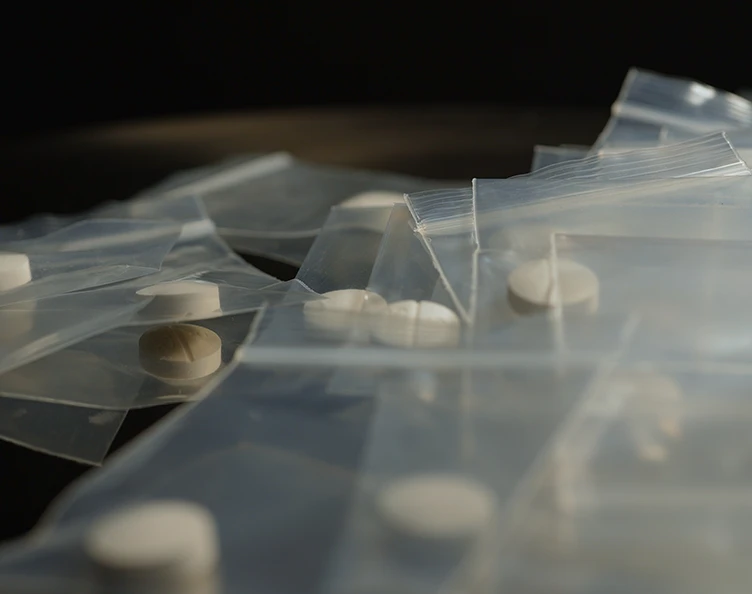 Multiple round, light-colored pills, some with a score, individually sealed in small, clear plastic zip-top bags and scattered on a dark, out-of-focus surface.